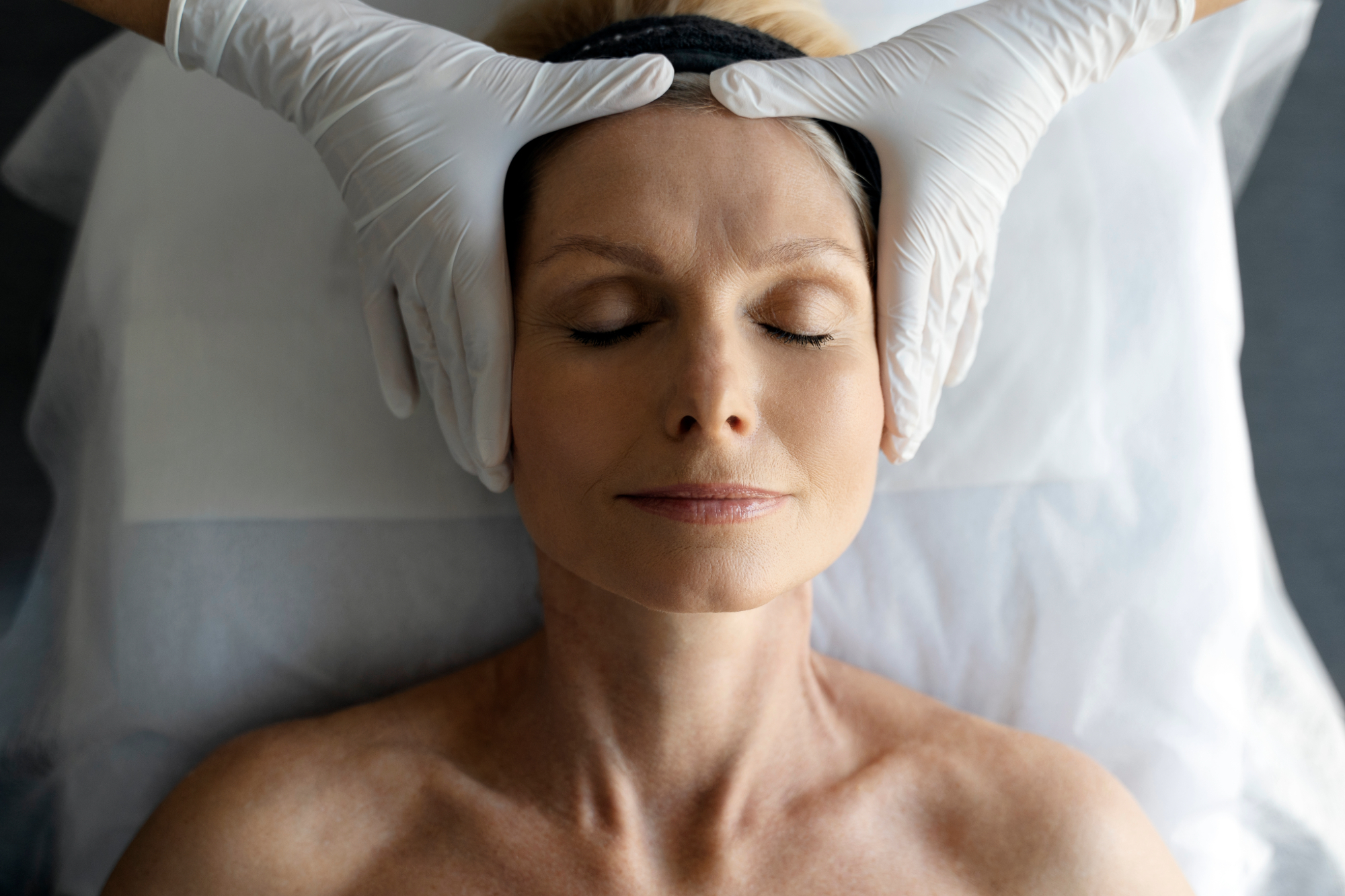Mature woman receiving a rejuvenating facial therapy with gloved hands gently positioned on her temples.